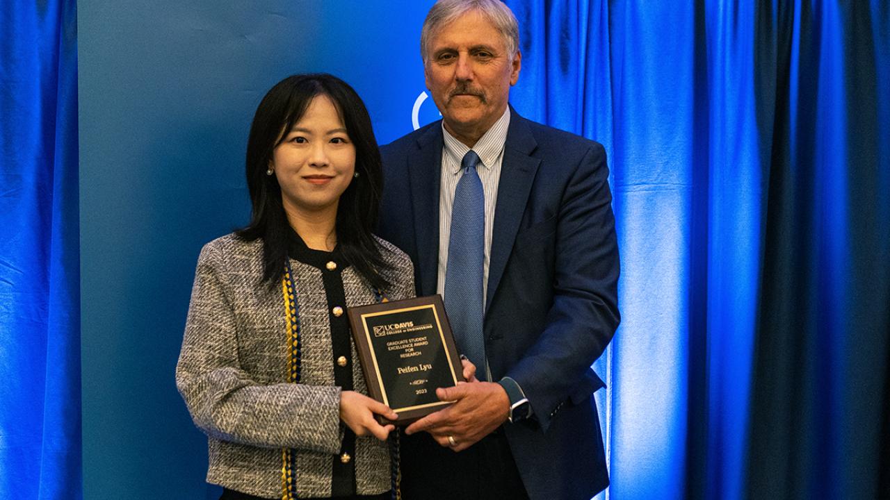 alt="Peifen Lyu stands to the left of Dean Richard Corsi, holding an award in front of a blue backdrop"